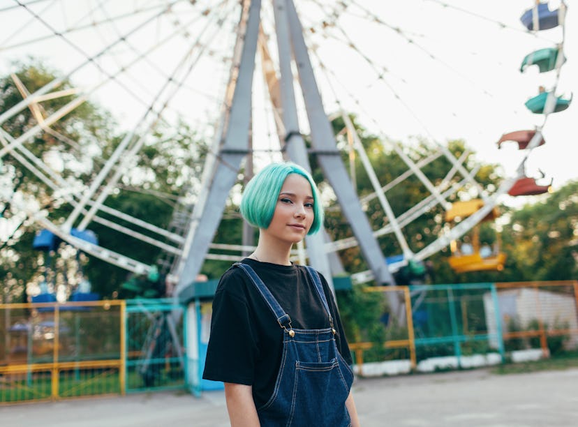 Teen girl with blue hair in at a fair in front of a Ferris wheel, preparing her Instagram with Ferri...