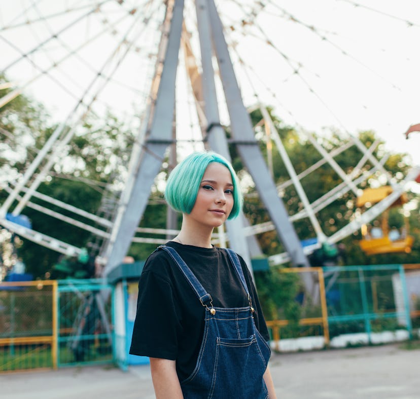 Teen girl with blue hair in at a fair in front of a Ferris wheel, preparing her Instagram with Ferri...