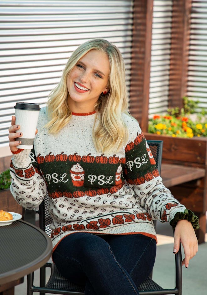 Woman sitting down, holding to-go coffee cup, wearing PSL sweater