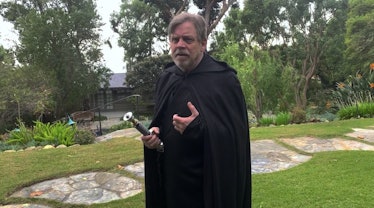 Mark Hamil holding a lightsaber while dressed in a black coat as Luke Skywalker