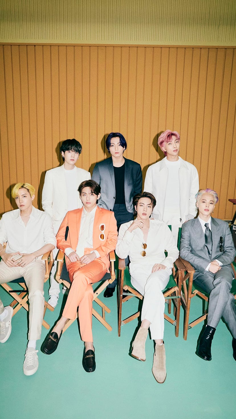 The members of BTS stand and sit in chairs as they gaze into the camera