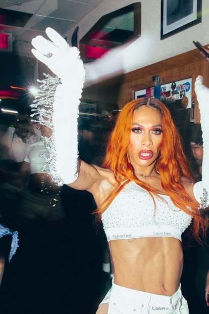A drag queen with red hair, wearing white gloves, white top and white bottoms