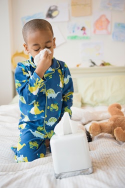 A Black boy wearing pajamas blows his nose with a tissue while sitting on a bed.