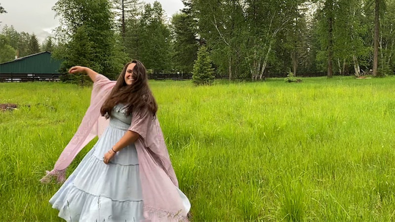 Emma Christensen in a blue dress and pink scarf dancing around a field next to the woods