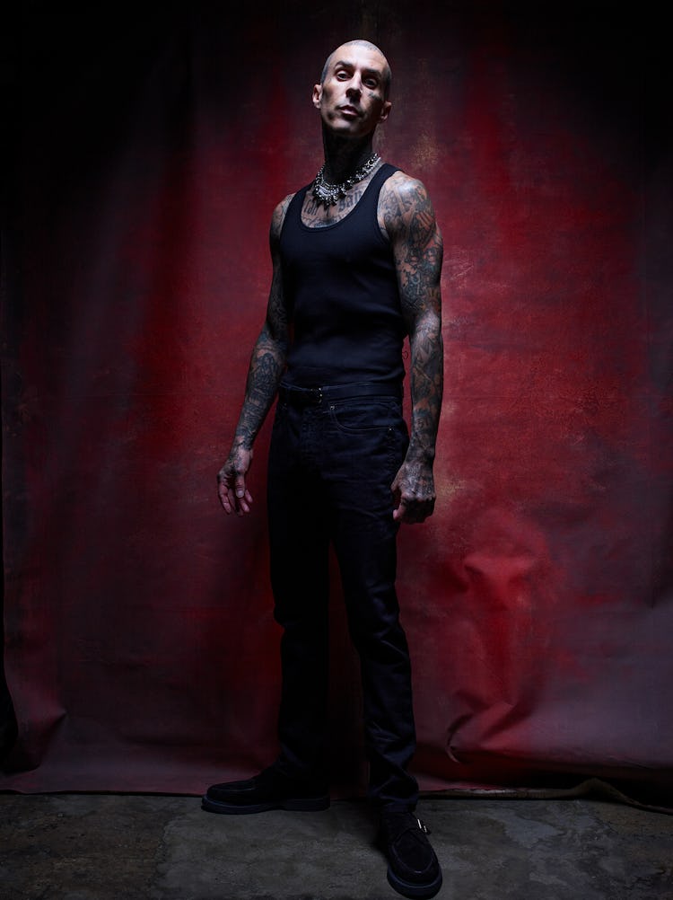 A full-length image of Travis Barker standing in front of a red backdrop.