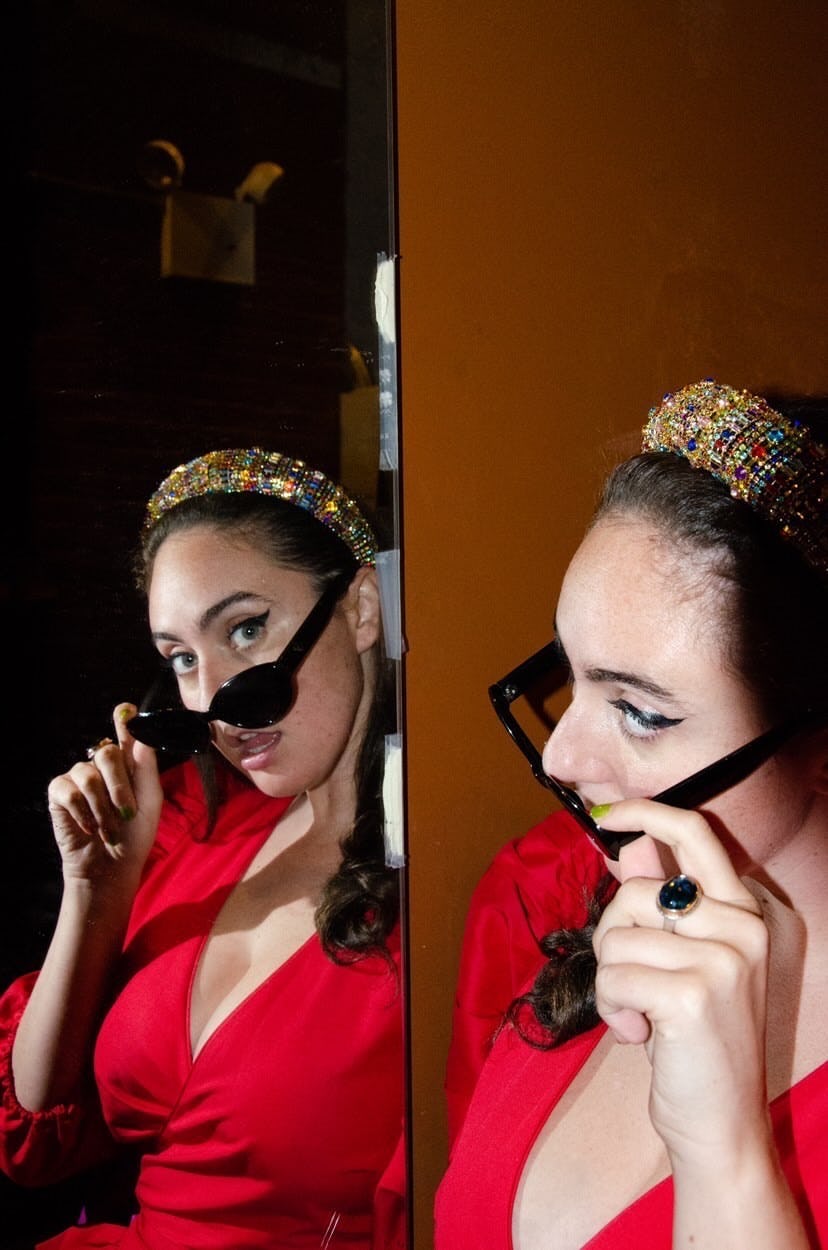 the comedian Catherine Cohen wearing a red dress and black sunglasses looking at herself in the mirr...