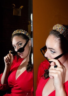 the comedian Catherine Cohen wearing a red dress and black sunglasses looking at herself in the mirr...