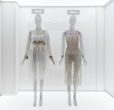 two short white dresses embellished with pressed and dried flowers on display in a museum case. 