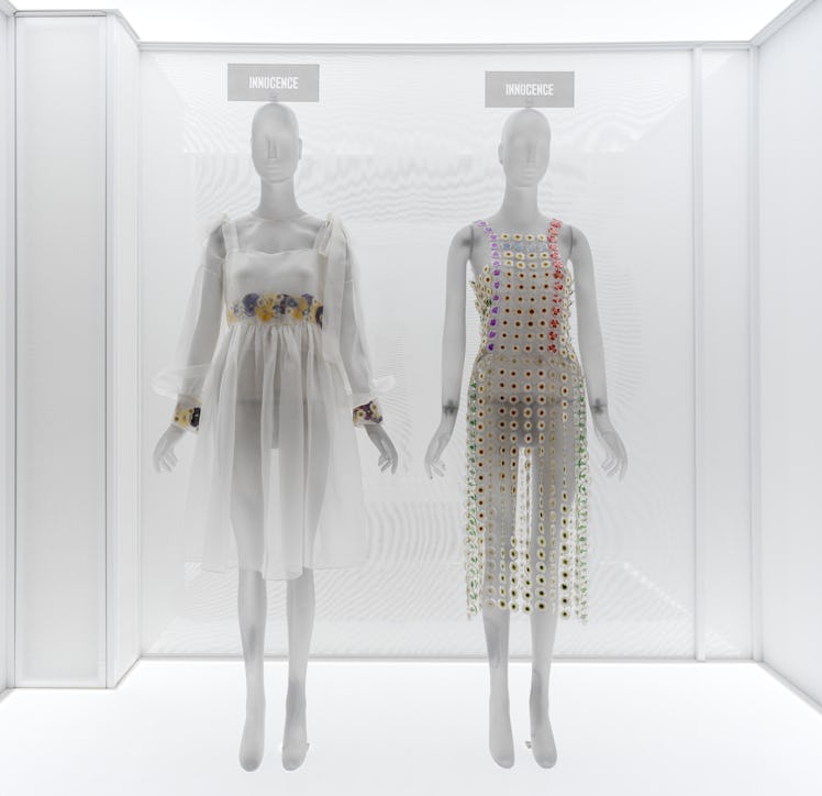 two short white dresses embellished with pressed and dried flowers on display in a museum case. 