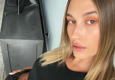 Hailey Bieber taking a selfie with minimalist makeup on, wearing a black crop top and pants