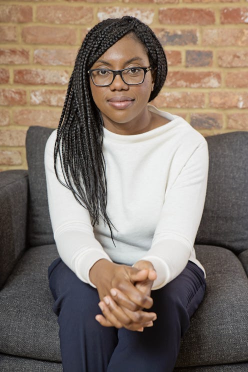 Kemi Badenoch sat on a grey sofa wearing a white jumper and blue smart trousers. Her hair is in brai...