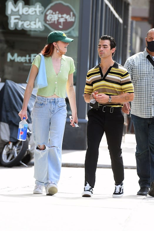 Sophie Turner wearing a green Jonas Brothers baseball hat, jeans, and a green top while carrying a b...