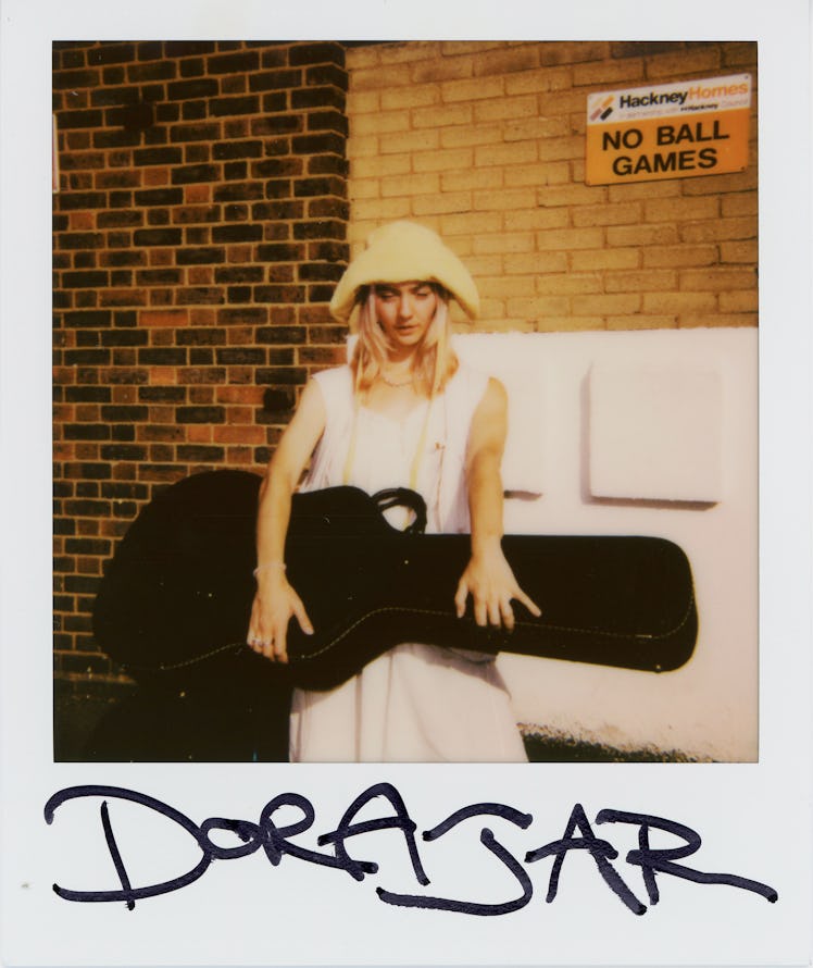 Polaroid of Gen Z Artist Dora Jar holding a guitar suitcase and wearing a pale pink dress with a bei...