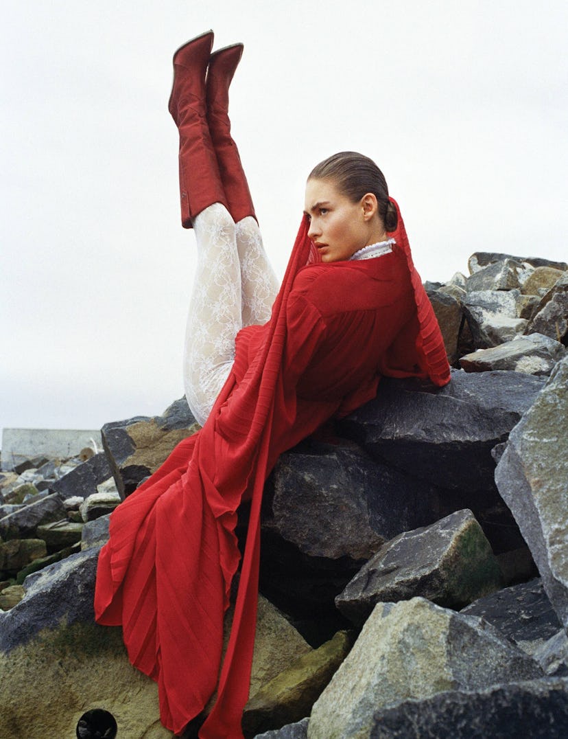 a woman in a red dress and tall brown boots reclines on a rocky outcropping