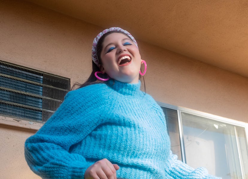 American comedian Meg Stalter wearing a blue sweater paired with a neon green skirt, and pink earrin...