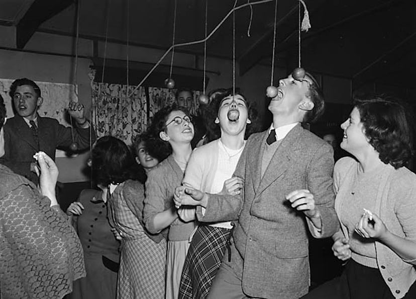 A group of party-goers bob for apples suspended from the ceiling.