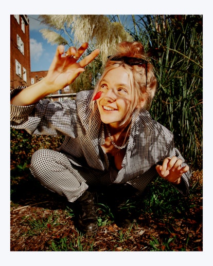 Gen Z artist Dora Jar crouching in the grass, smiling, wearing a checkered pattern suit