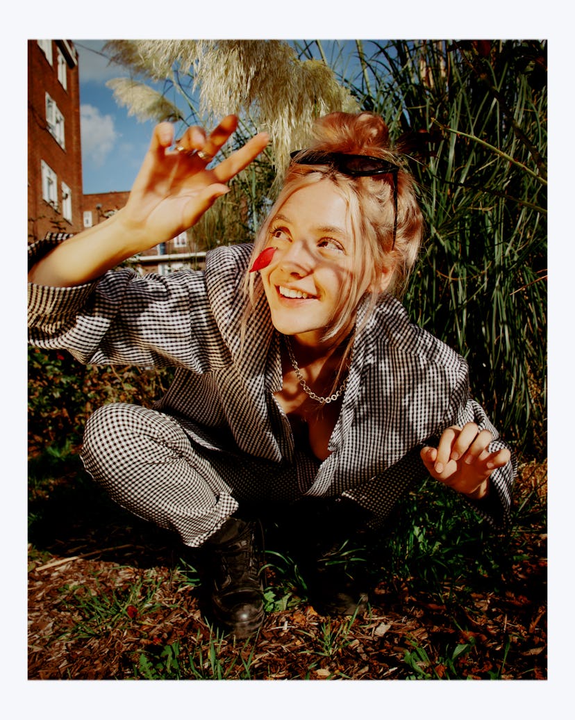 Gen Z artist Dora Jar crouching in the grass, smiling, wearing a checkered pattern suit