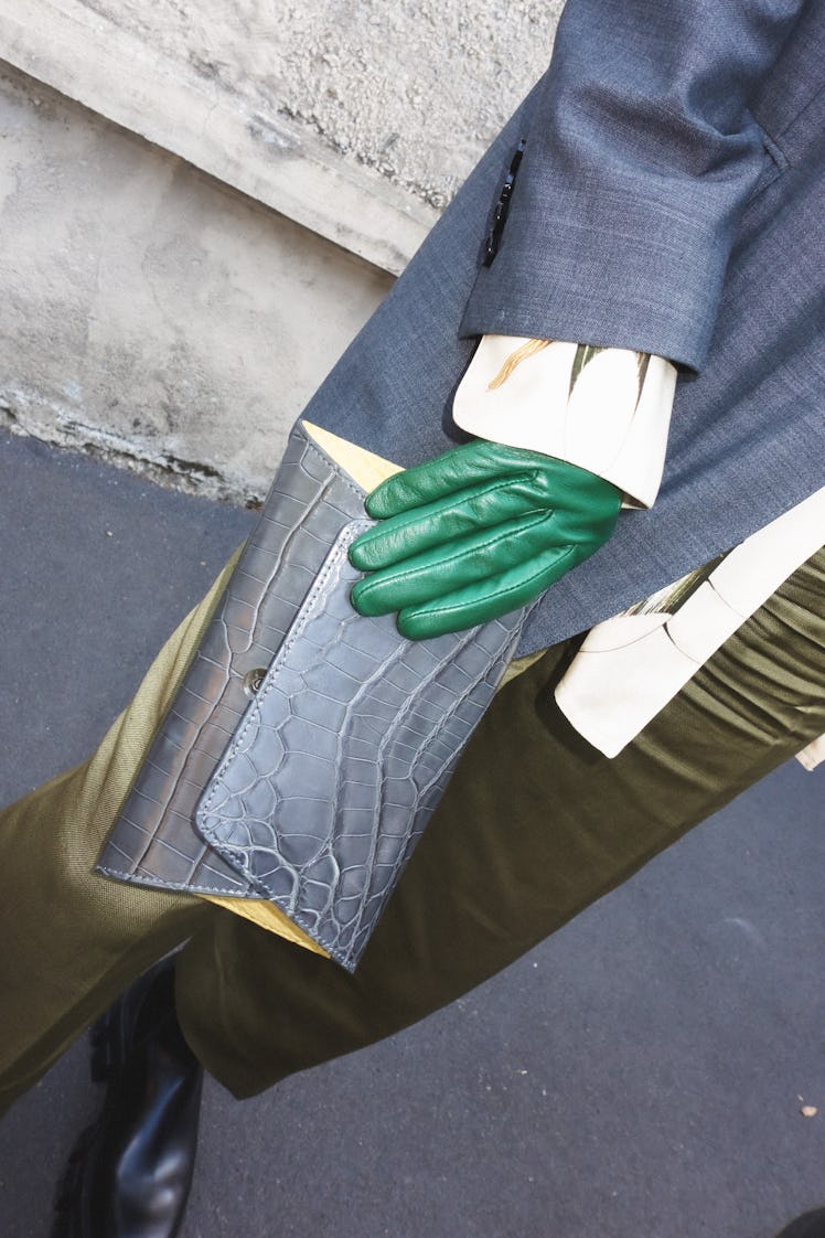 A close-up of a woman in a grey coat and beige trousers, green gloves and a grey bag
