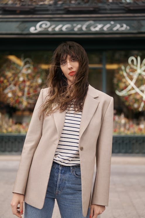 A brunette in a striped shirt, jeans and a beige blazer wearing a red lipstick in the French way
