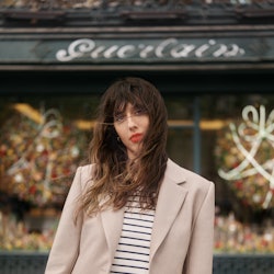 A brunette in a striped shirt, jeans and a beige blazer wearing a red lipstick in the French way