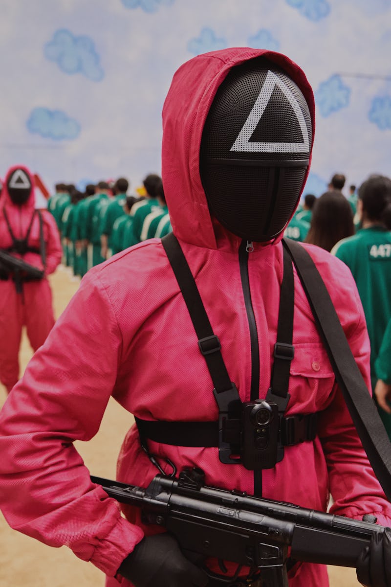 A character in a pink uniform in Squid Games walking among the people in green tracksuits 