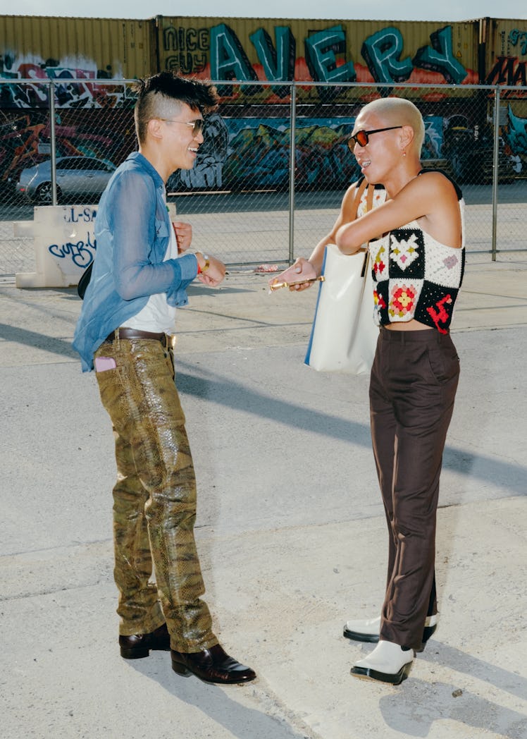 Two friends speak on the street. Both wear tight pants and shirts.