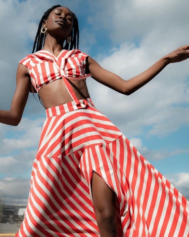 A black model wearing a red and white striped top and skirt set in front of a blue sky with clouds