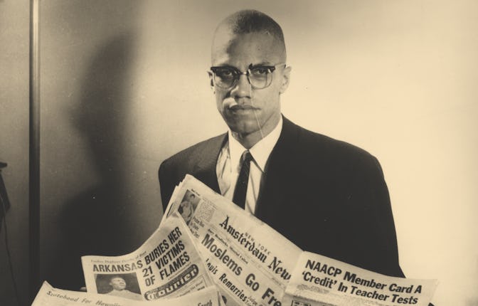 Portrait of human rights activist Malcolm X reading stories about himself in a pile of newspapers, c...