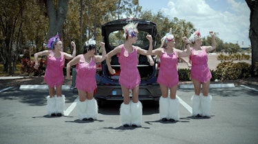 Five women wearing pink dresses and white fury boots while dancing in front of a car in the "Calenda...
