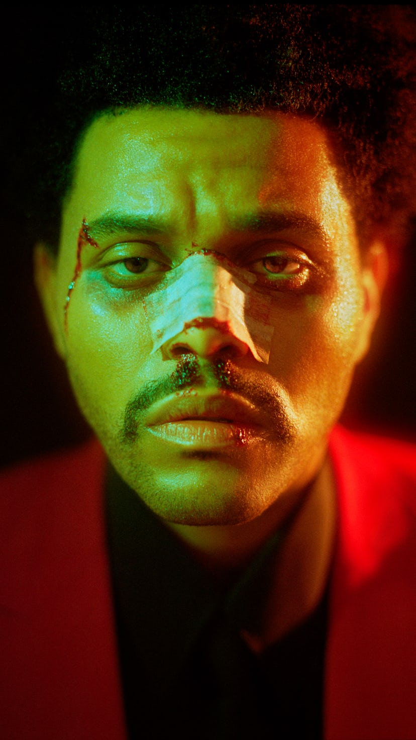 The Weeknd wears a black shirt, a red suit, and a patch on his bloody nose