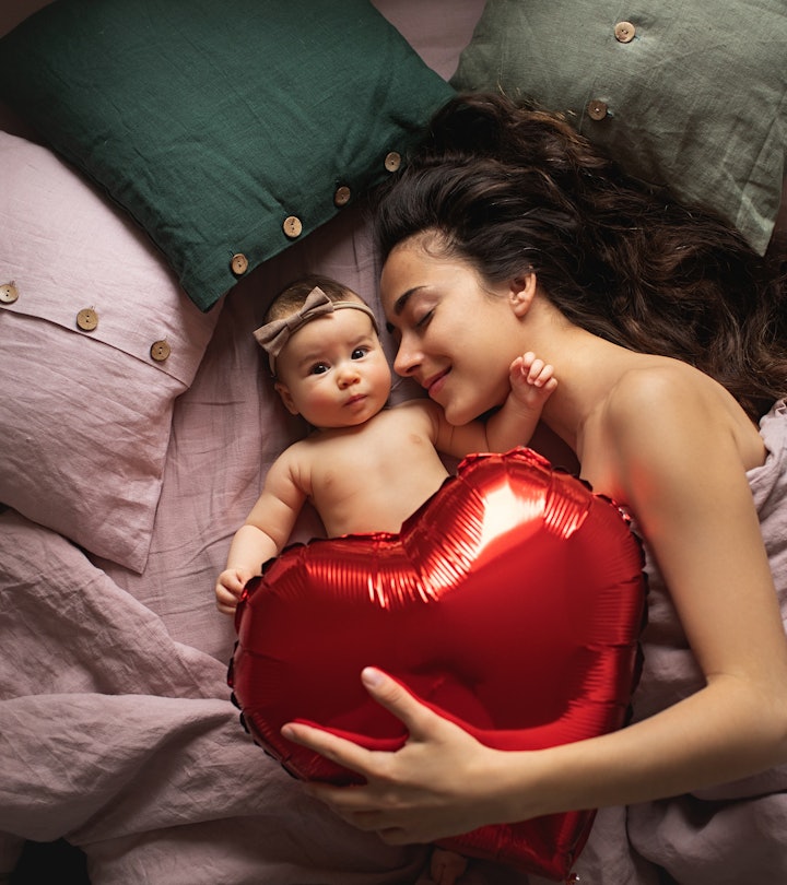 mom with newborn lying in bed, holding a red heart balloon 