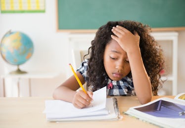 A kid at a school desk writes on paper while pouting.