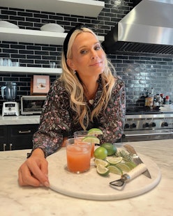 A blonde lady posing in her kitchen with two orange cocktail glasses and lime slices in front of her