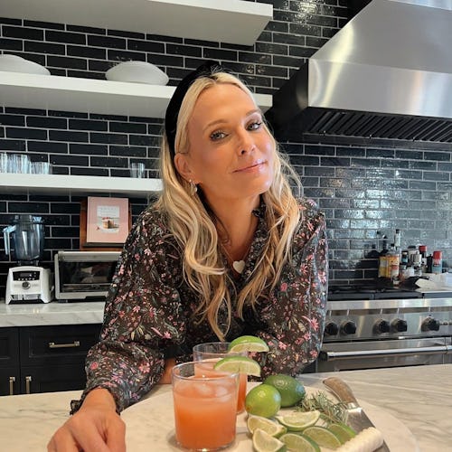 A blonde lady posing in her kitchen with two orange cocktail glasses and lime slices in front of her
