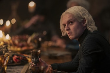 Aegon II Targaryen sitting at the dining table while holding a glass of wine in his hand