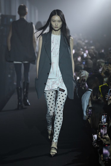 A model walking in a black blazer, white mini dress, and white leggings with black dots at the Raf S...