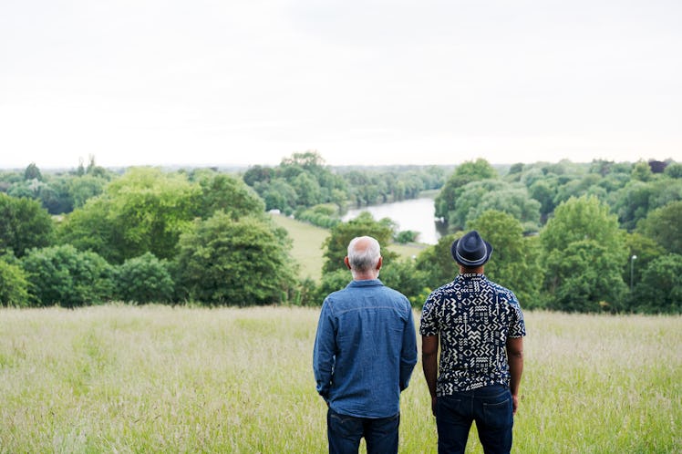 Two men in a field, looking into the distance: one in a short-sleeved shirt and fedora and the other...