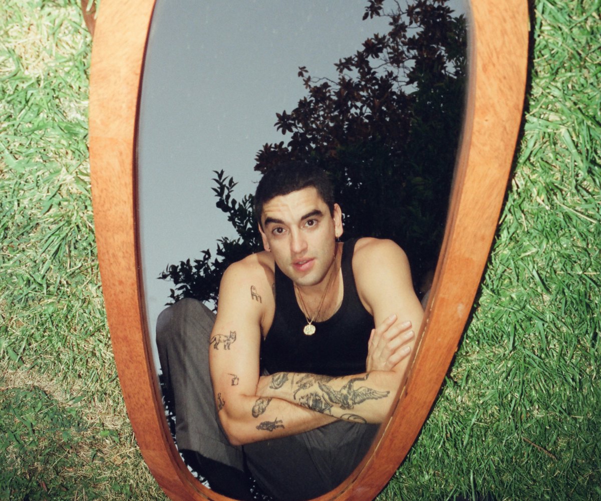 Adam Melchor squatting in front of an oval mirror with a light-wood frame which is placed in the gra...