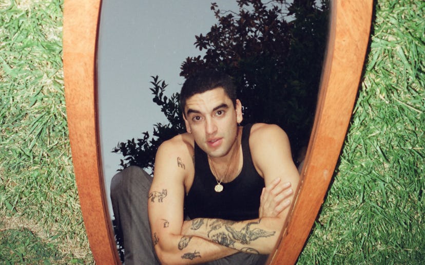 Adam Melchor squatting in front of an oval mirror with a light-wood frame which is placed in the gra...