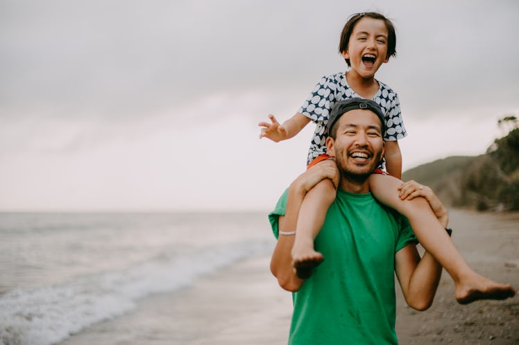 A laughing dad carries his daughter on his shoulders at the beach.