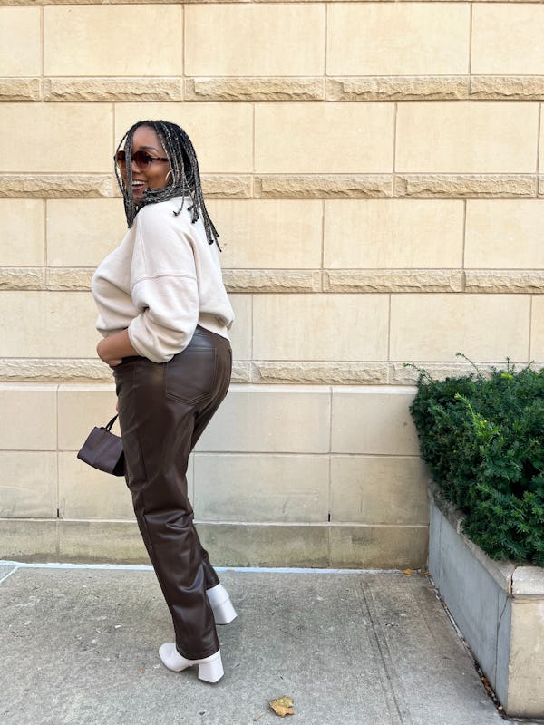 A mid-size model posing in a white sweater, brown vegan leather pants, white heel boots, and a brown…