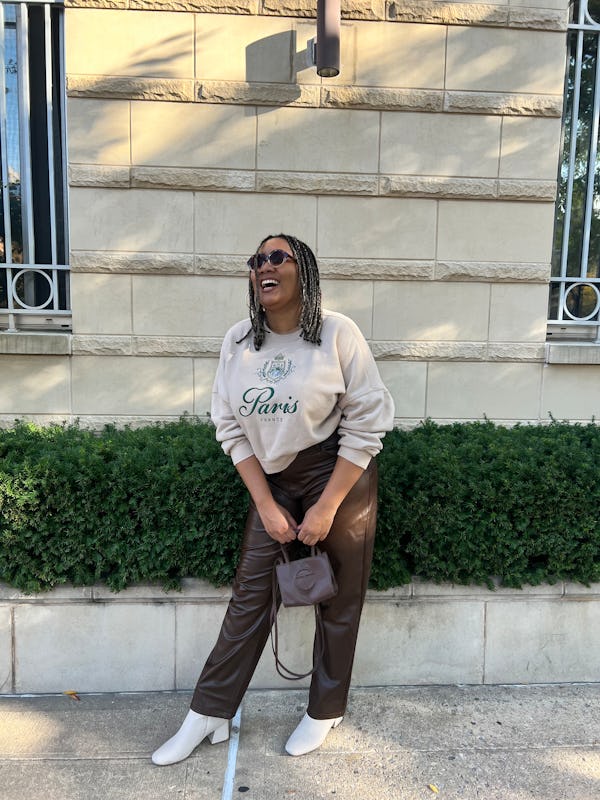 A mid-size model posing in a white sweater, brown vegan leather pants, white heel boots, and a brow…