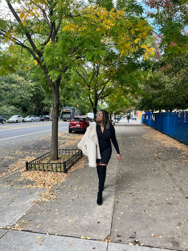 A mid-size woman walking in a black long-sleeve shirt dress and holding white shearling jacket on he…