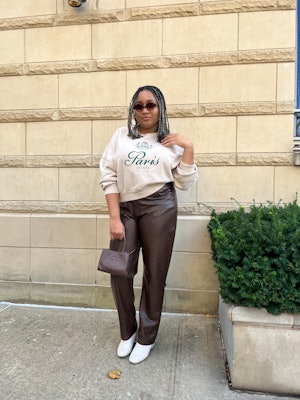 A mid-size model posing in a white sweater, brown vegan leather pants, white heel boots, and a brown...