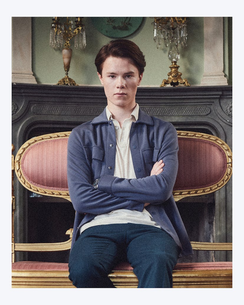 A portrait of ‘Young Royals’ star Edvin Ryding sitting on a royal burgundy & gold bench in a collare...