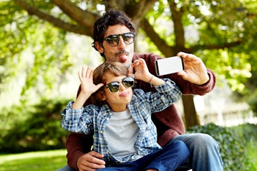 A dad and son sitting on a park bench wearing sunglasses and making silly faces while taking a selfi...