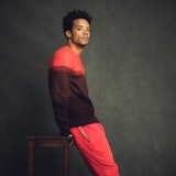 A portrait of Jacob Anderson wearing a knit red and burgundy sweater and red pants, leaning on a sto...