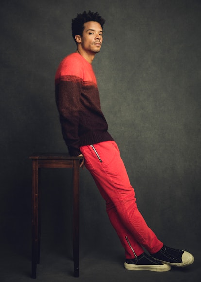 A portrait of Jacob Anderson wearing a knit red and burgundy sweater and red pants, leaning on a sto...