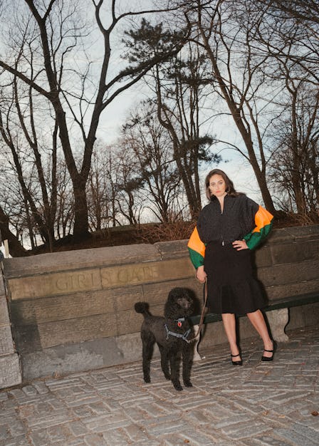 Catherine Cohen standing in front of Central Park in the fall with a black dog